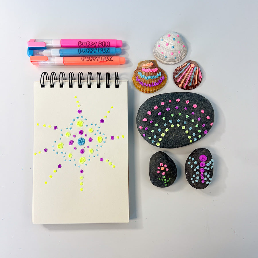 3 seashells decorated with lines and dots, three black rocks decorated with dots to form a rainbow and flower and a sketchpad with dot decoration that all are using OOLY Magic puffy pens, three puffy pens pink blue and orange are in frame.