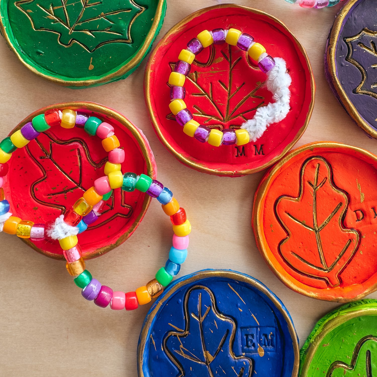 Colorful beaded bracelets placed on top of clay dishes with leaf designs, painted in red, green, orange, and blue with gold accents, surrounded by Paintology acrylic paint tubes. OOLY Craft blog