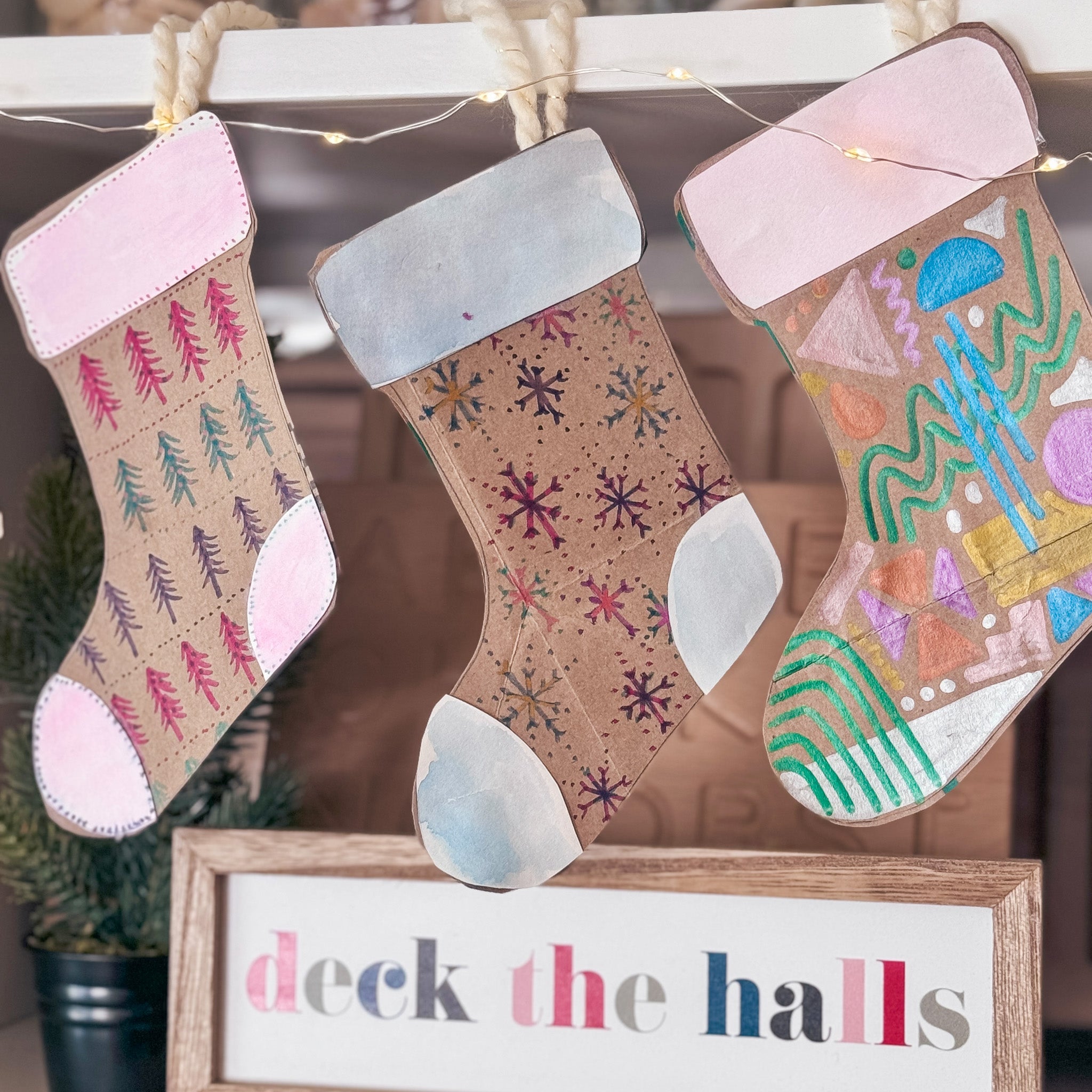 Completed upcycled paper bag stockings with designed with trees, snowflakes and geometric designs hanging from a mantle with a sign that says deck the halls.