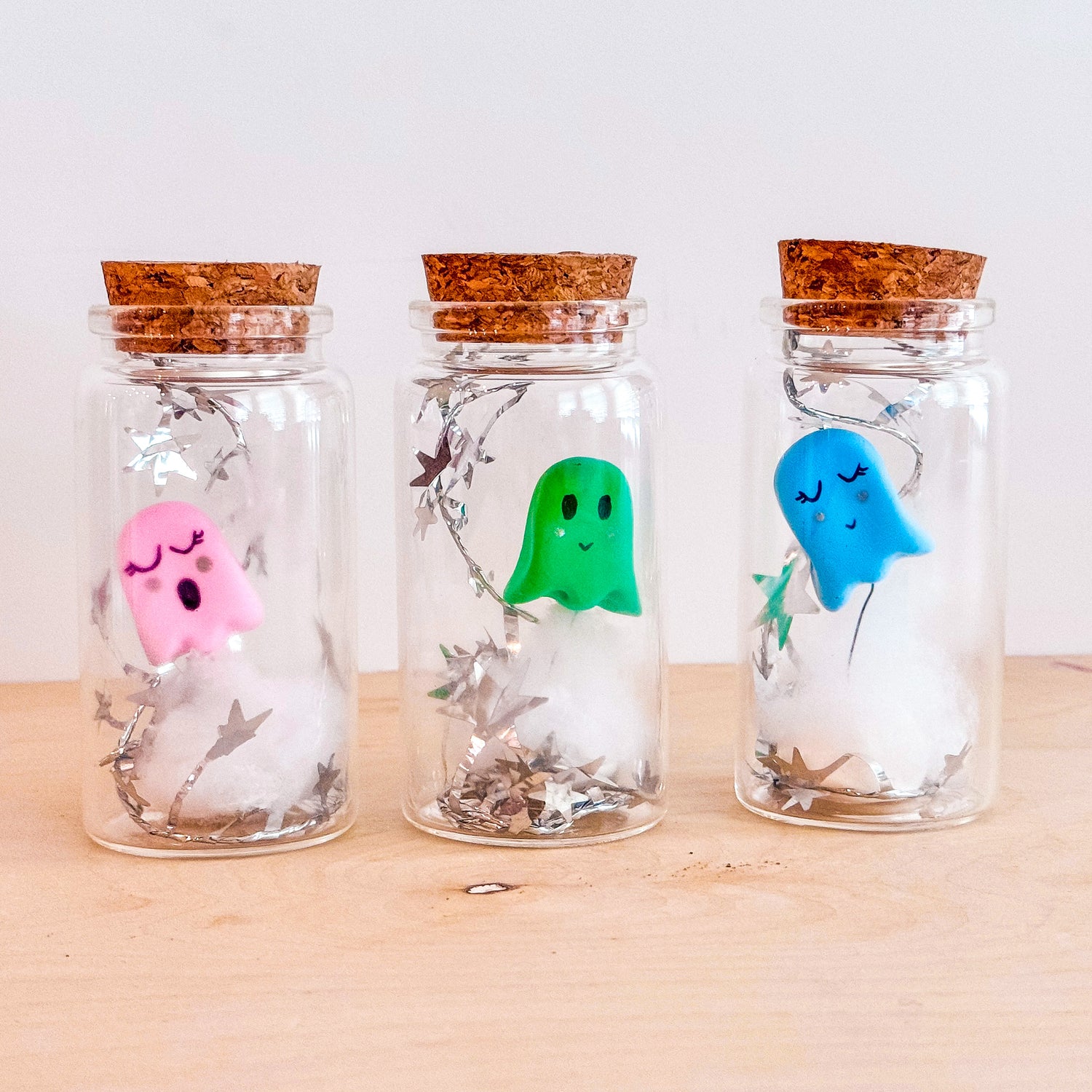 Three glass bottles with cork lids, each containing a brightly colored ghost made from OOLY Creatibles air dry clay, floating above cotton stuffing with star-shaped garland inside. OOLY Craft blog.