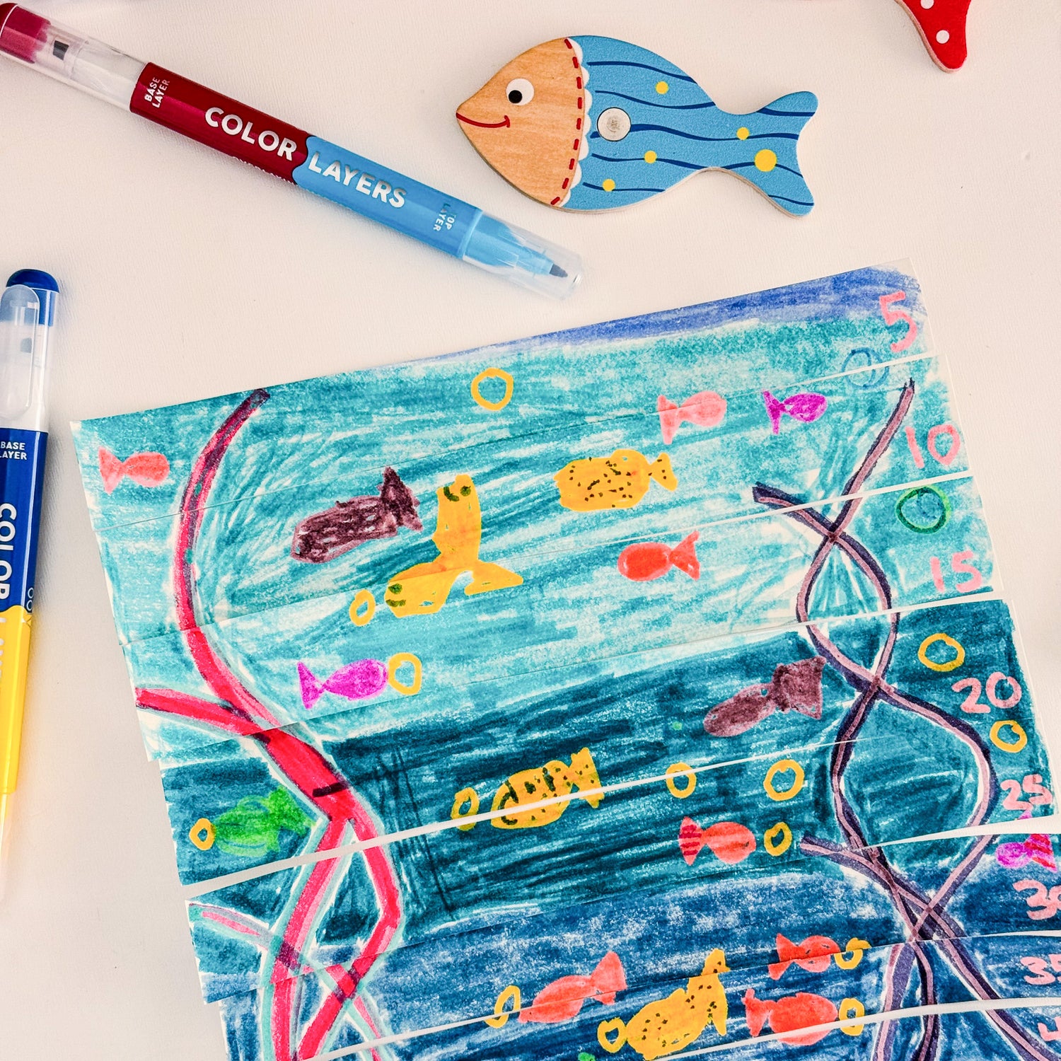 Drawing of an Ocean theme using OOLY Color Layers Markers with fish and coral, cut into strips like a puzze, next to a Color Layers Marker and a cute fish decoration