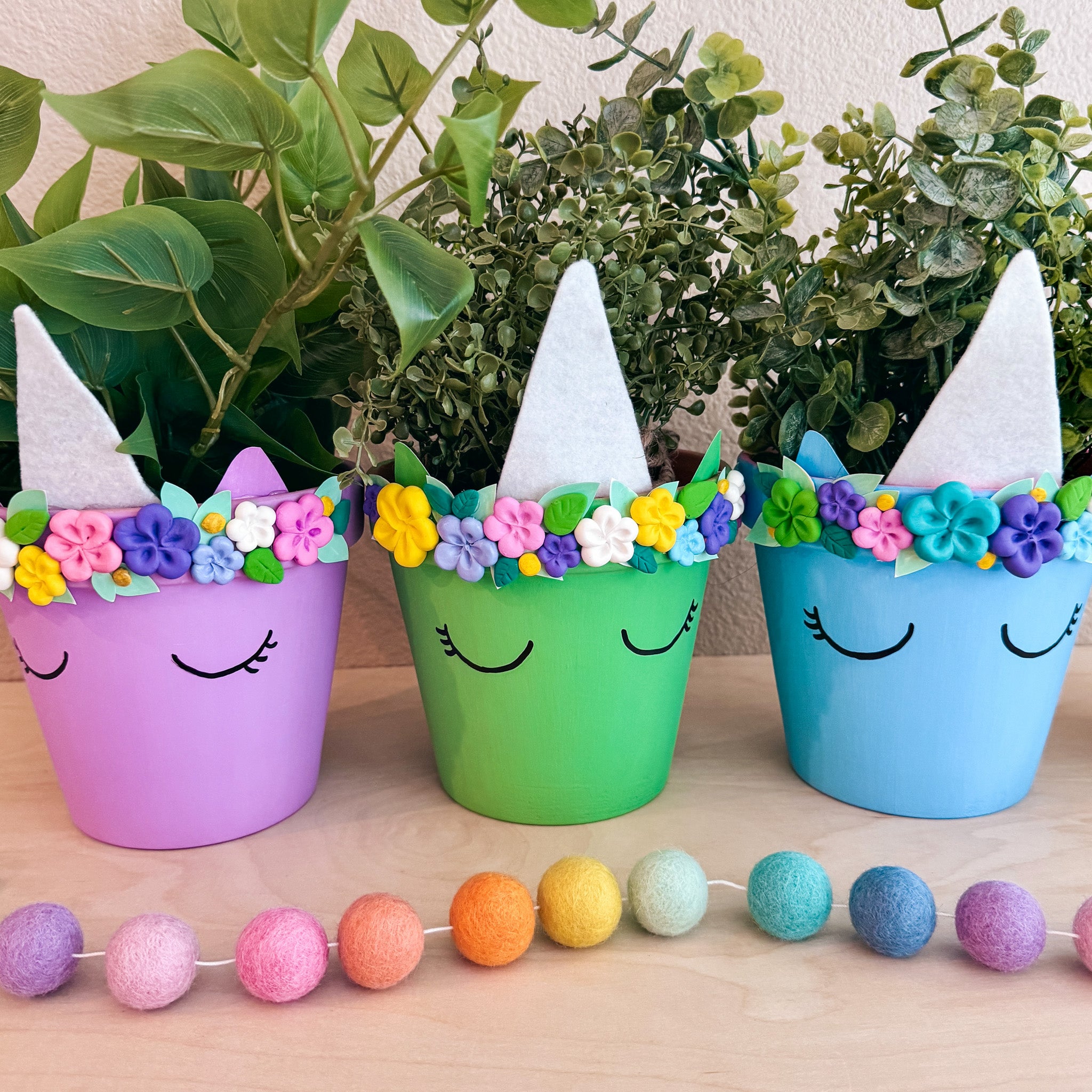 Three pastel-colored DIY unicorn planters on a table with flower crowns made from air-dry clay.