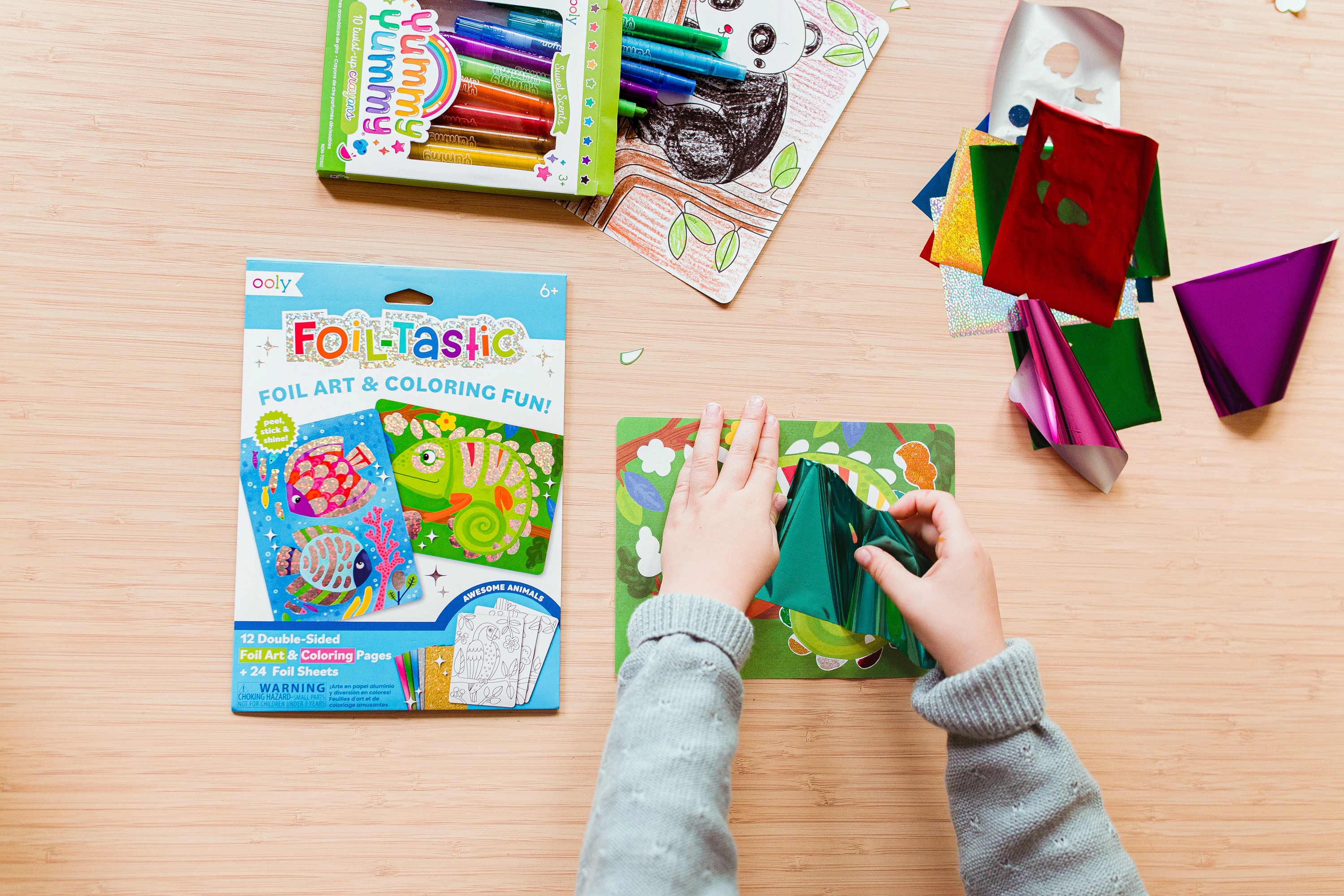 Girl putting foil on OOLY Foil-Tastic Foil Art Coloring Set with set of Yummy Crayons on wooden table