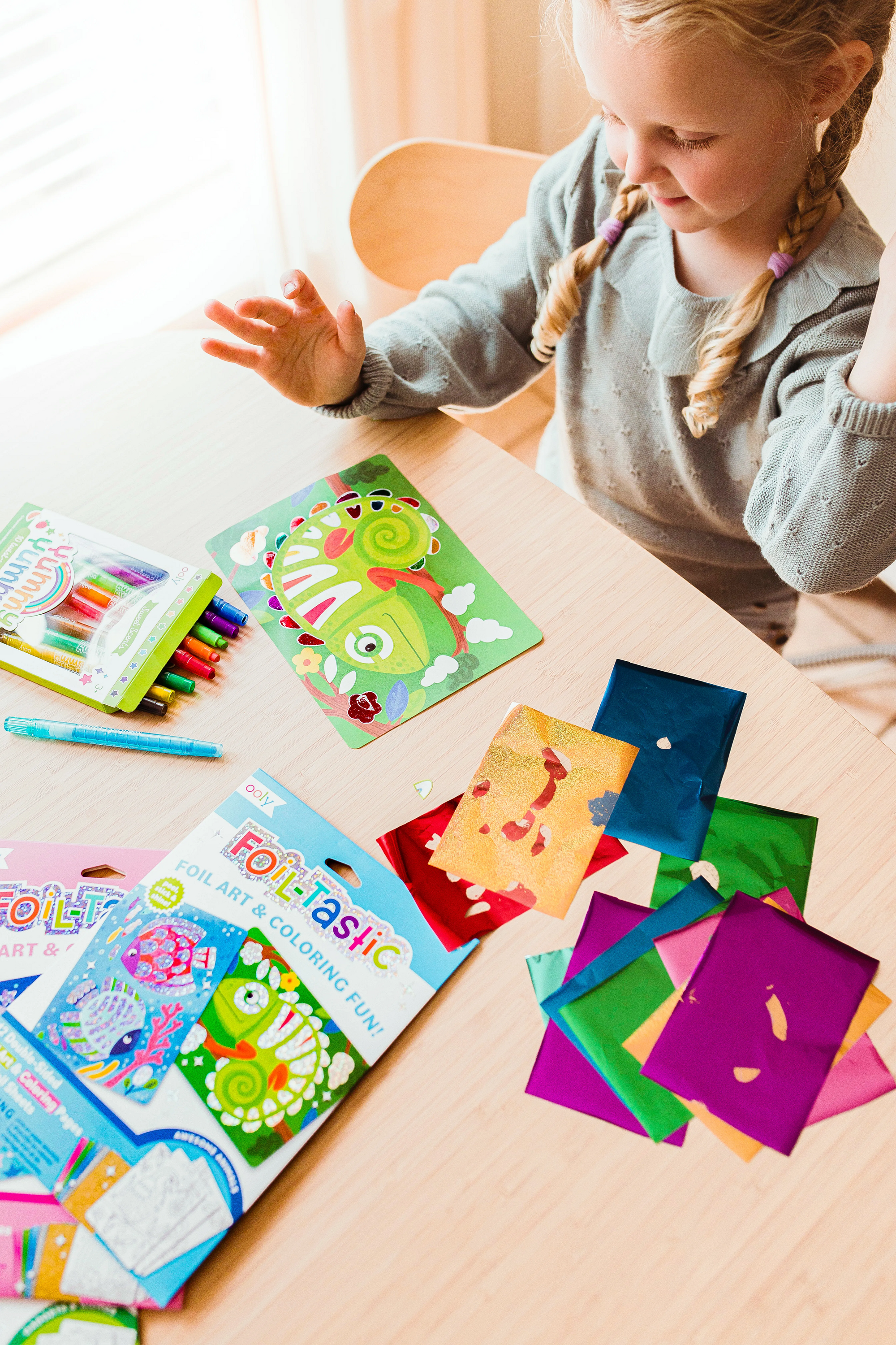 Little girl looking at chamileon art from OOLY Foil-Tastic Foil Art Coloring Set with set of Yummy Crayons on wooden table