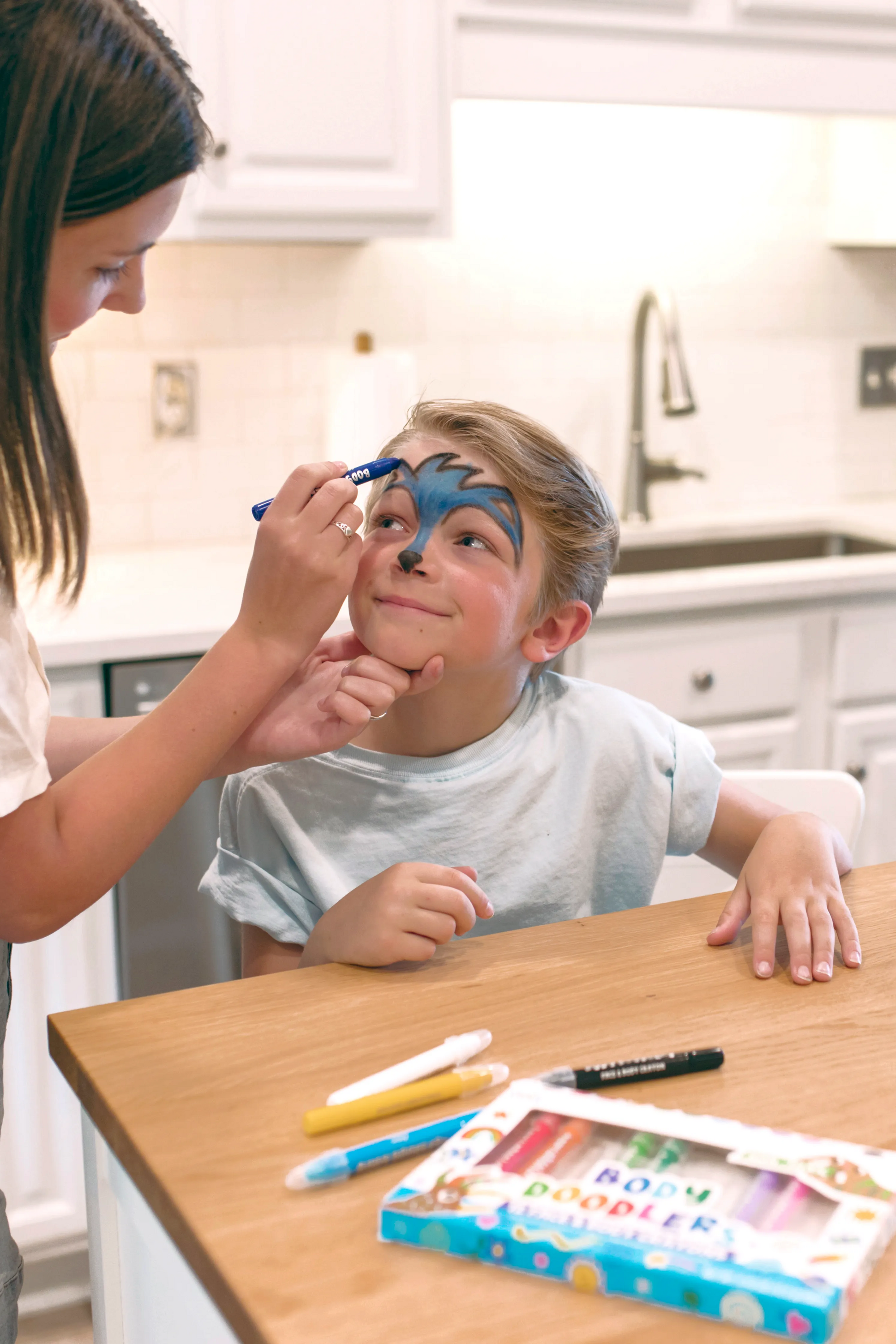 Woman drawing blue with OOLY Body Doodlers - Face & Body Crayon on little boy's face