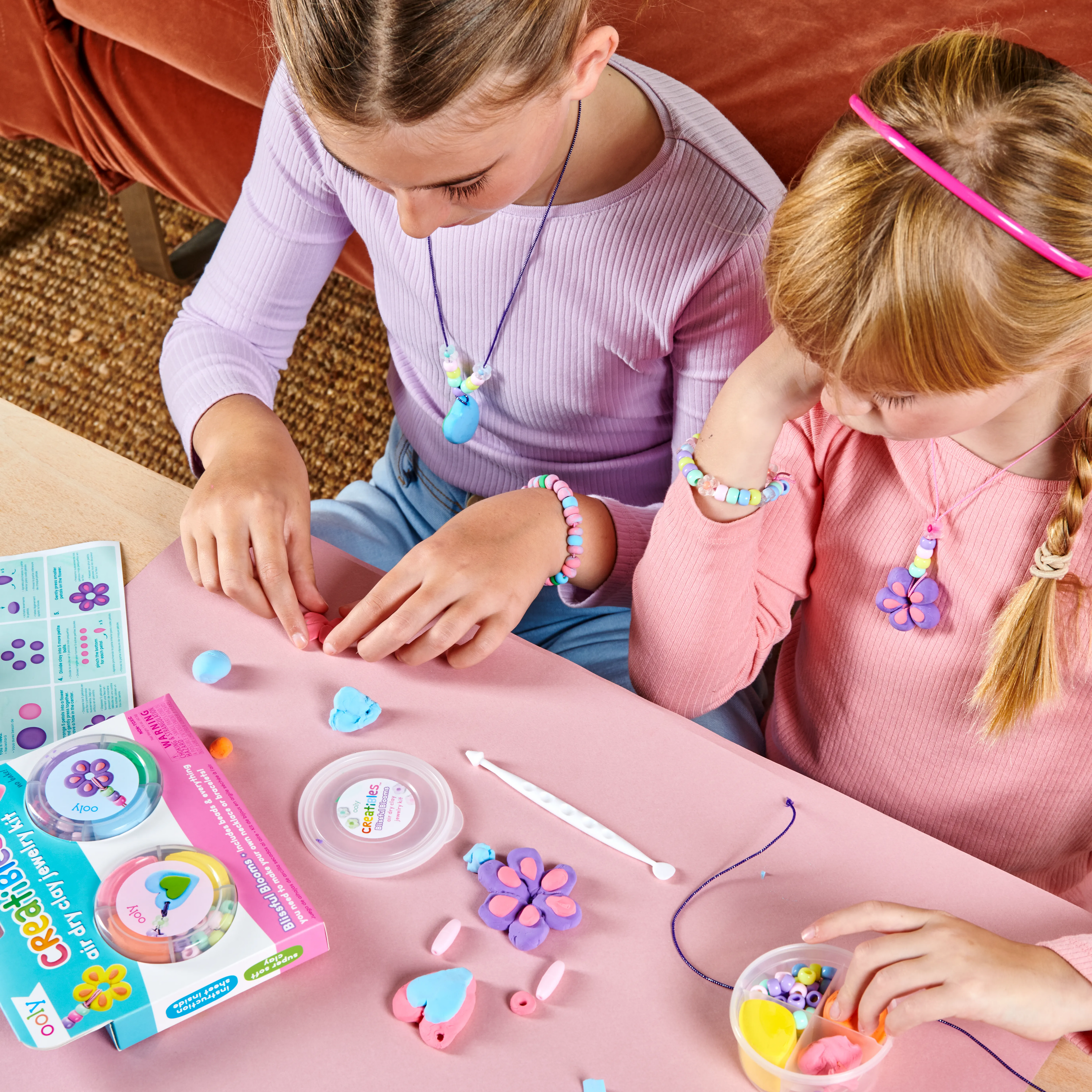 Two girls crafting jewelry from the OOLY Creatibles Air Dry Clay Jewelry Kit - Blissful Blooms set on a pink table top 