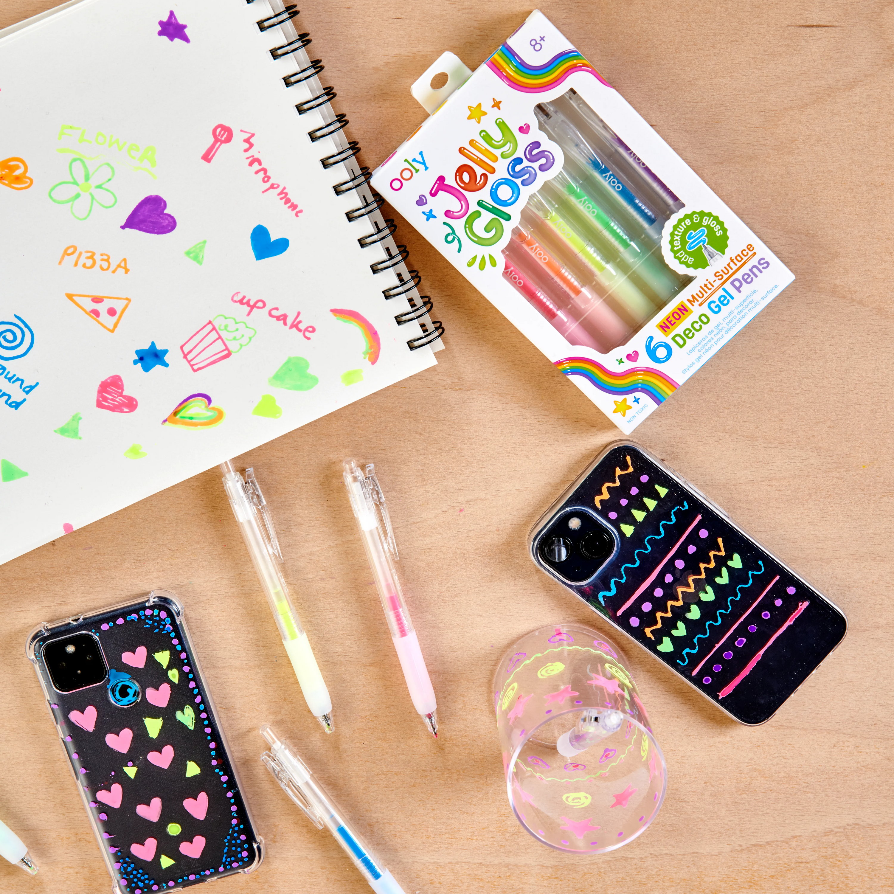 A table top setting with decorated phones, a sketchbook and pens from a set of OOLY Jelly Gloss Neon Multi-Surface Deco Gel Pens