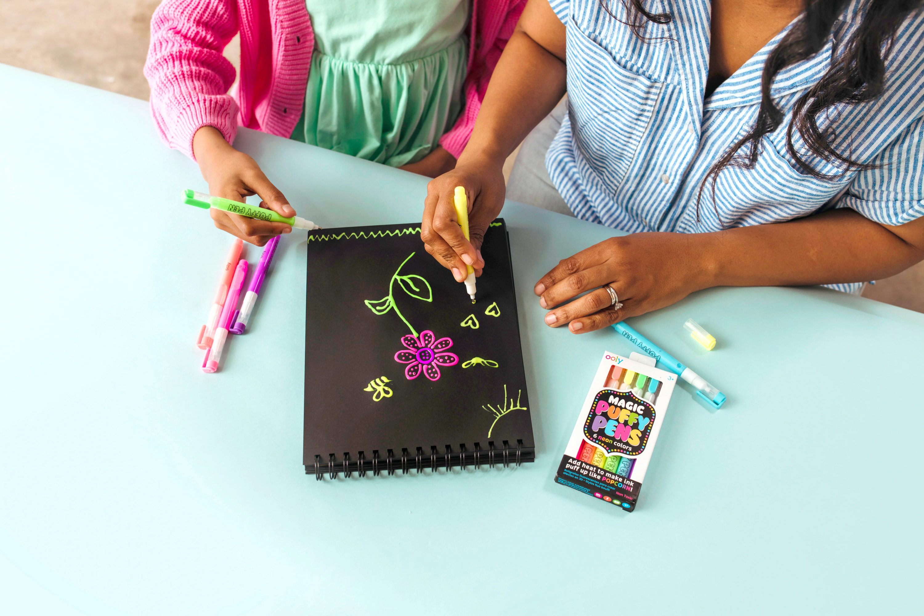 Two people painting on black sketchpad with OOLY Magic Puffy Neon Pens Painting Pad