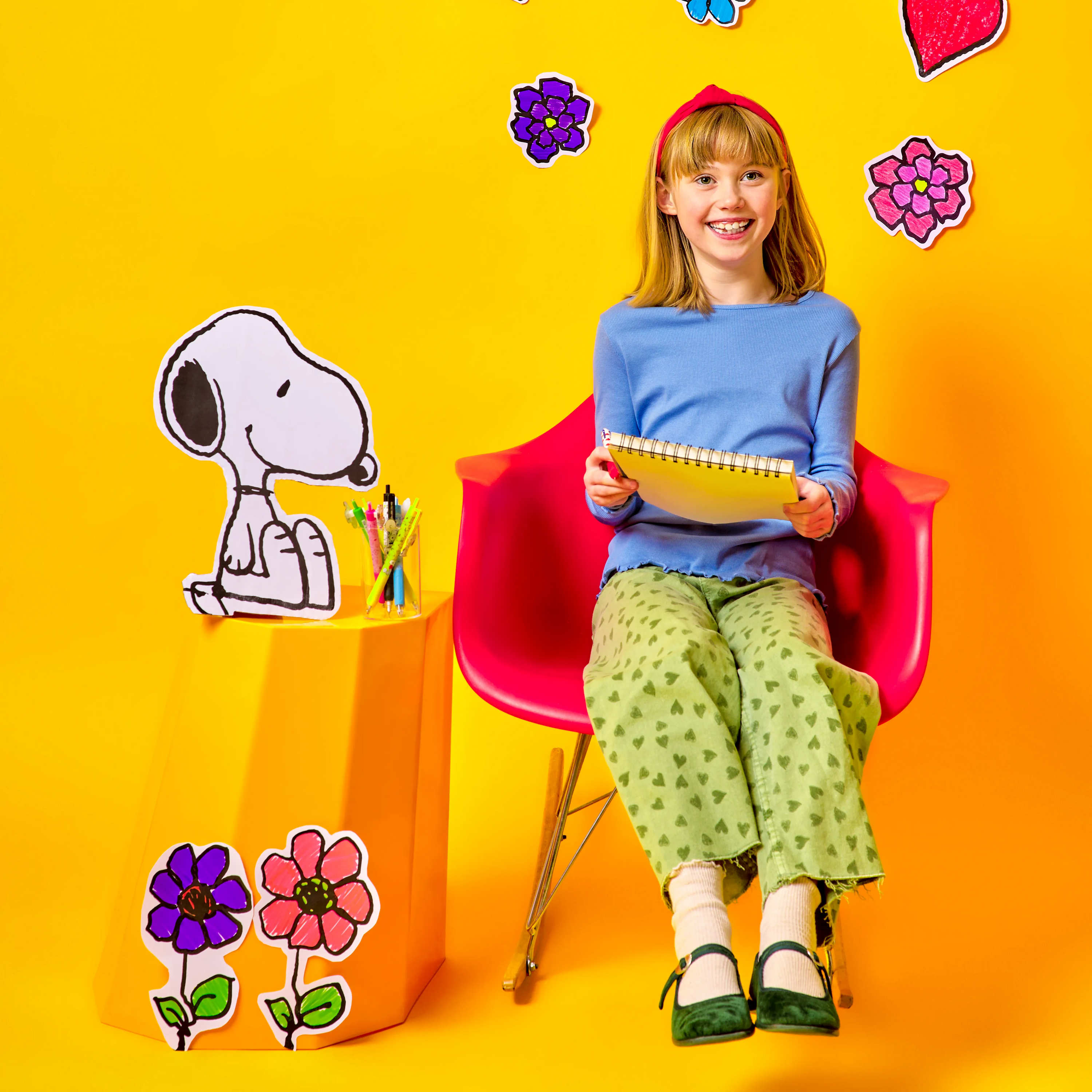 Girl in yellow background holding sketchbook and sitting in a chair with Snoopy cutout and Peanuts writing pens