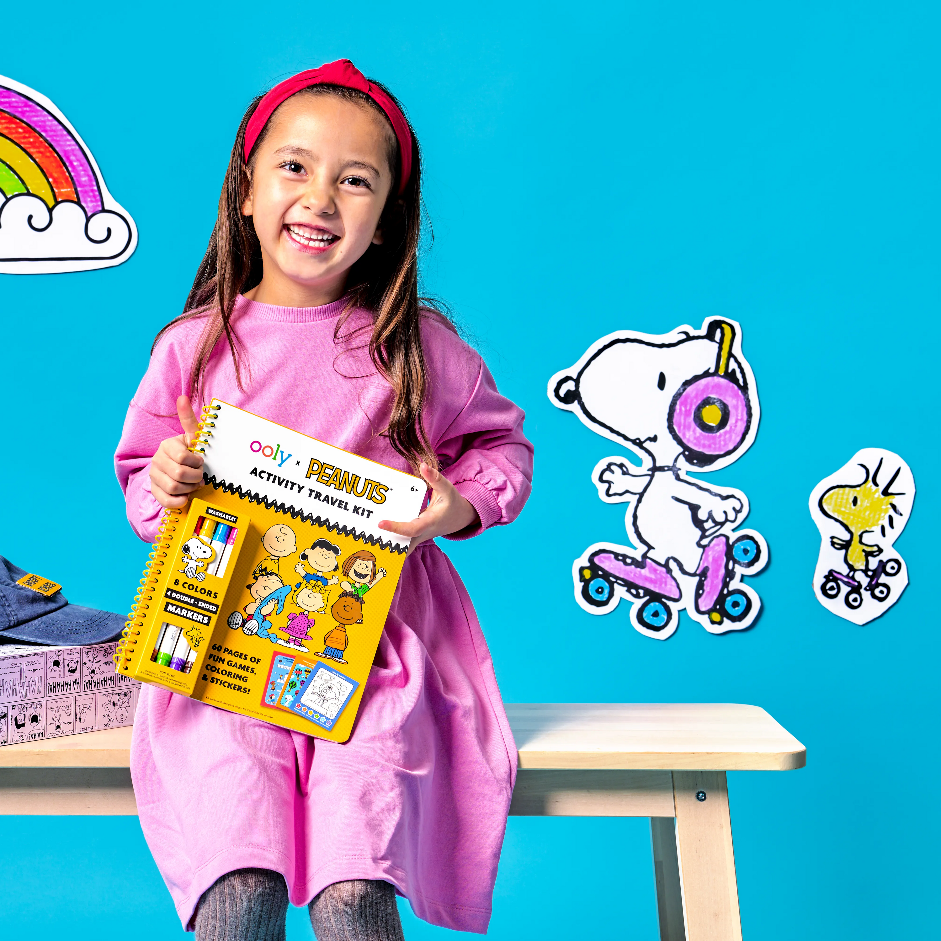 Girl smiling while holding a OOLY x Peanuts Activity Travel Kit & Double-Ended Markers set on blue background with Snoopy and Woodstock skating