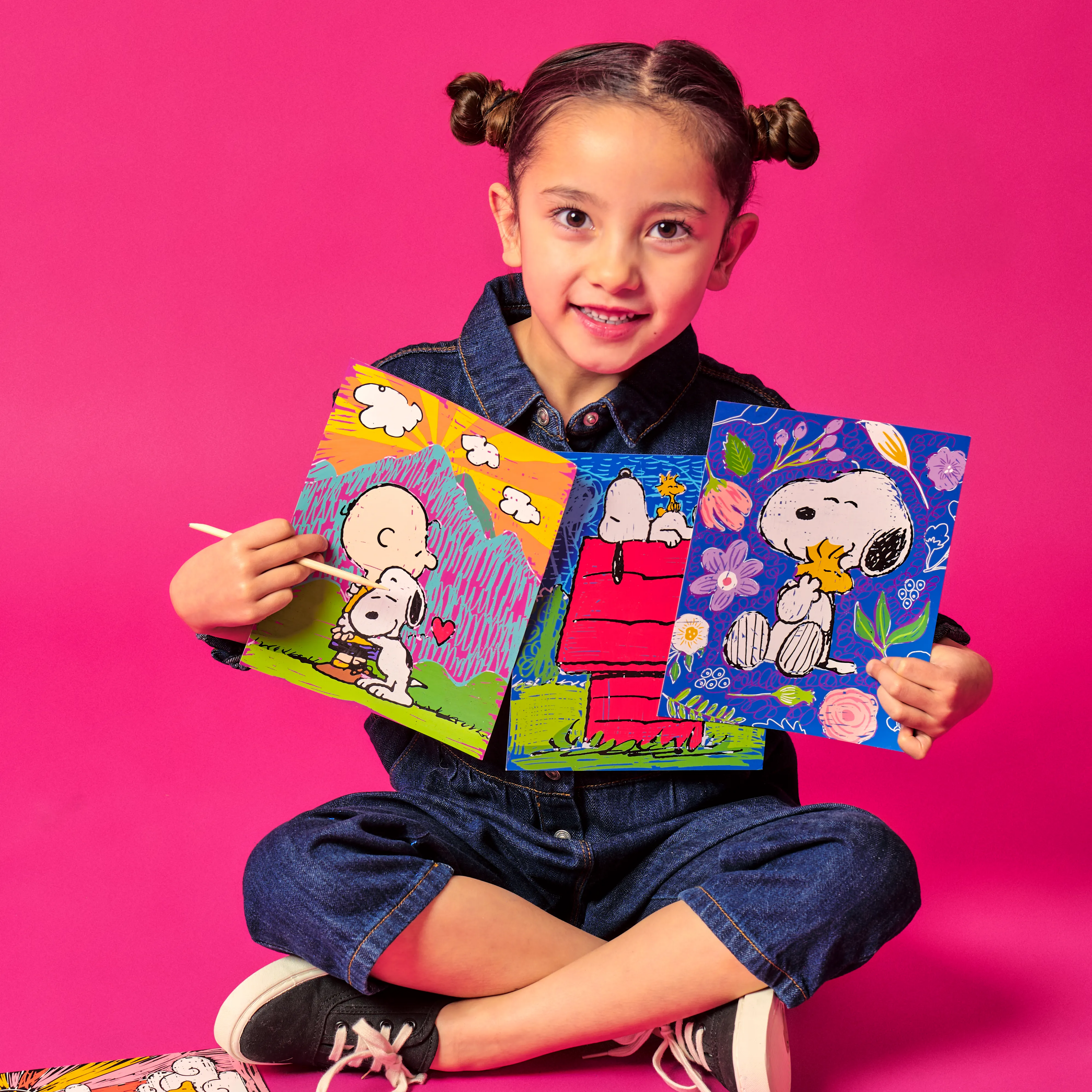 Girl holding up scratch art cards of Charlie Brown and Snoopy from the OOLY x Peanuts Scratch Art Kit