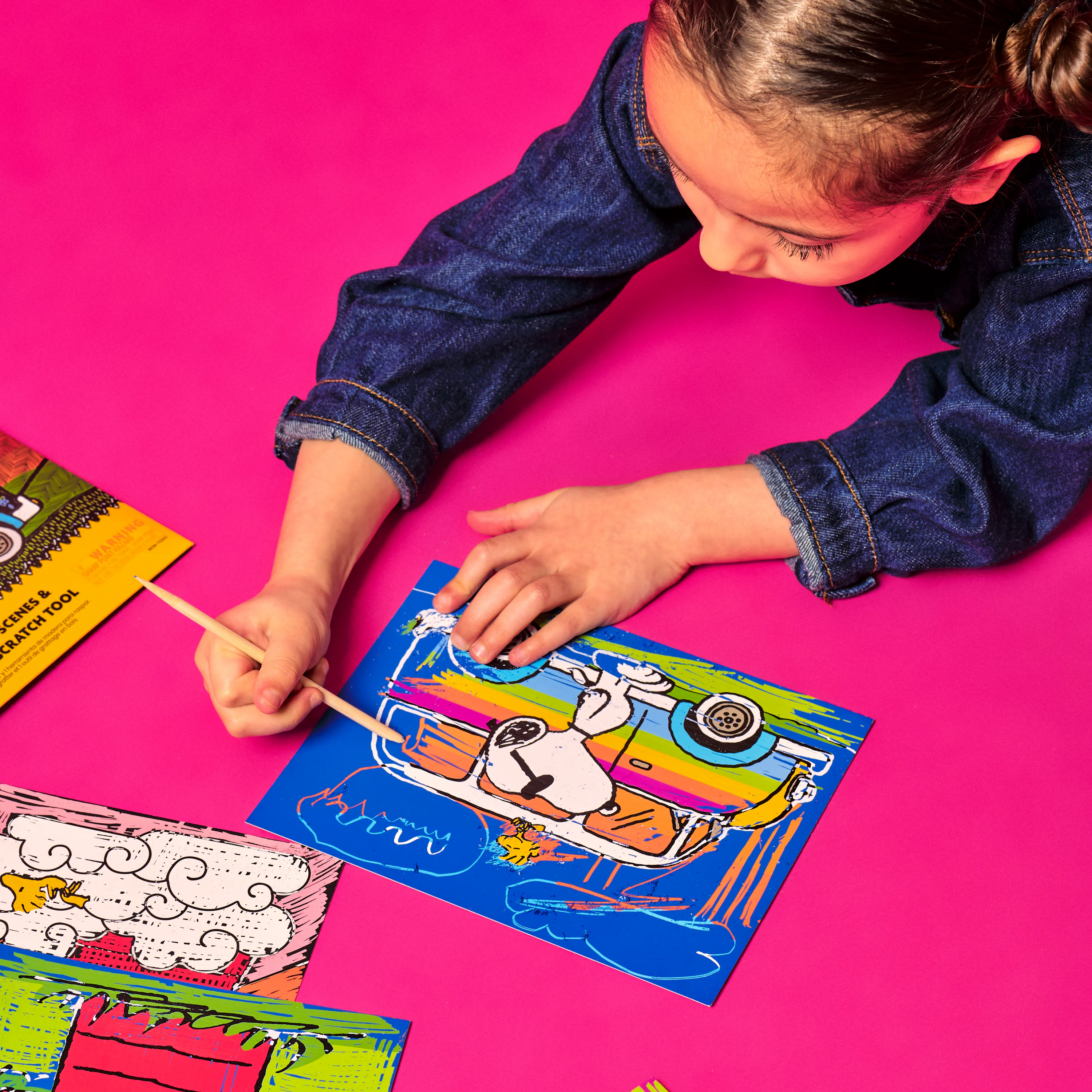 Girl scratching card with Snoopy and rainbow colored car from the OOLY x Peanuts Scratch Art Kit