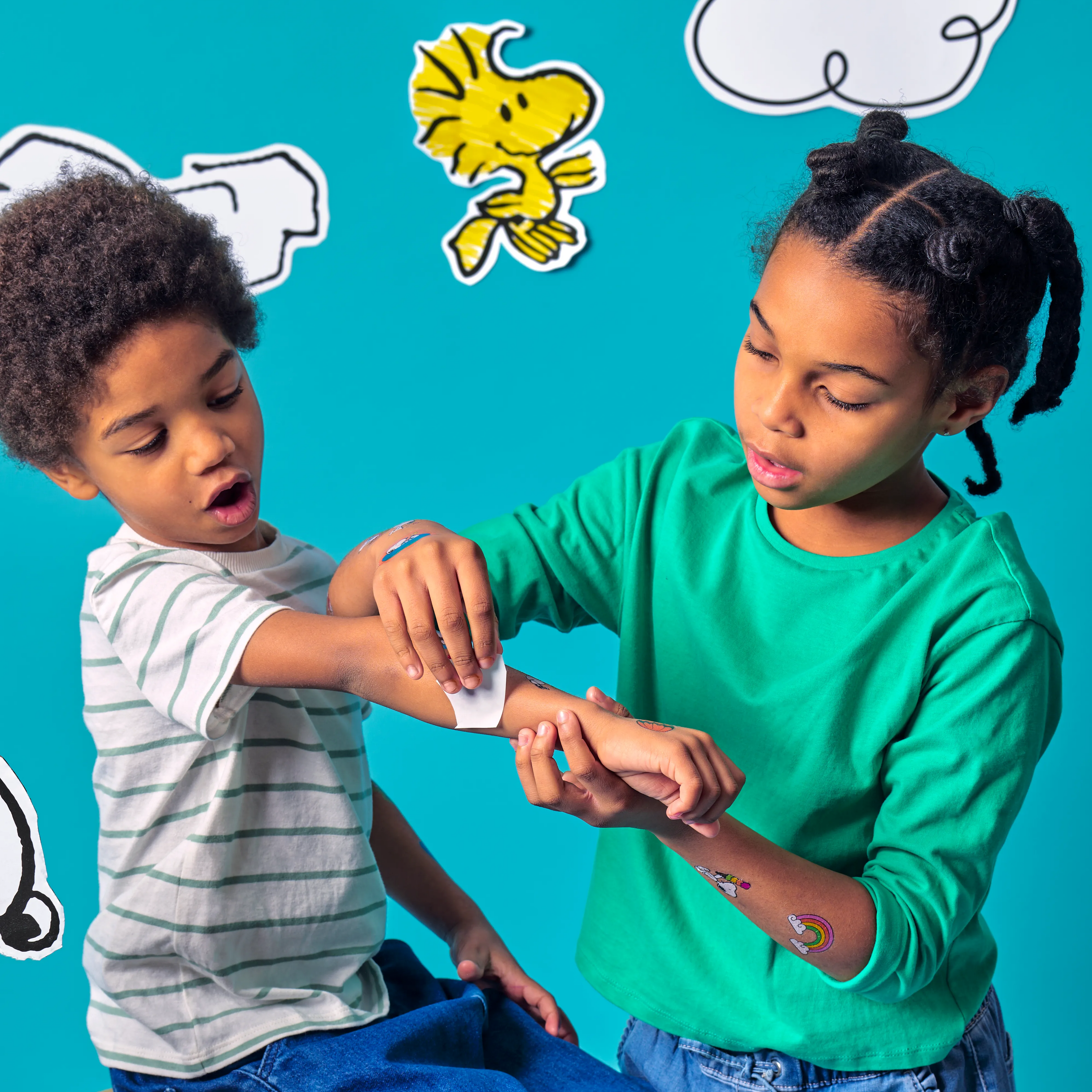 Two kids against a cloudy blue background with Woodstock applying temporary Tattoos from an OOLY x Peanuts Temporary Tattoos set