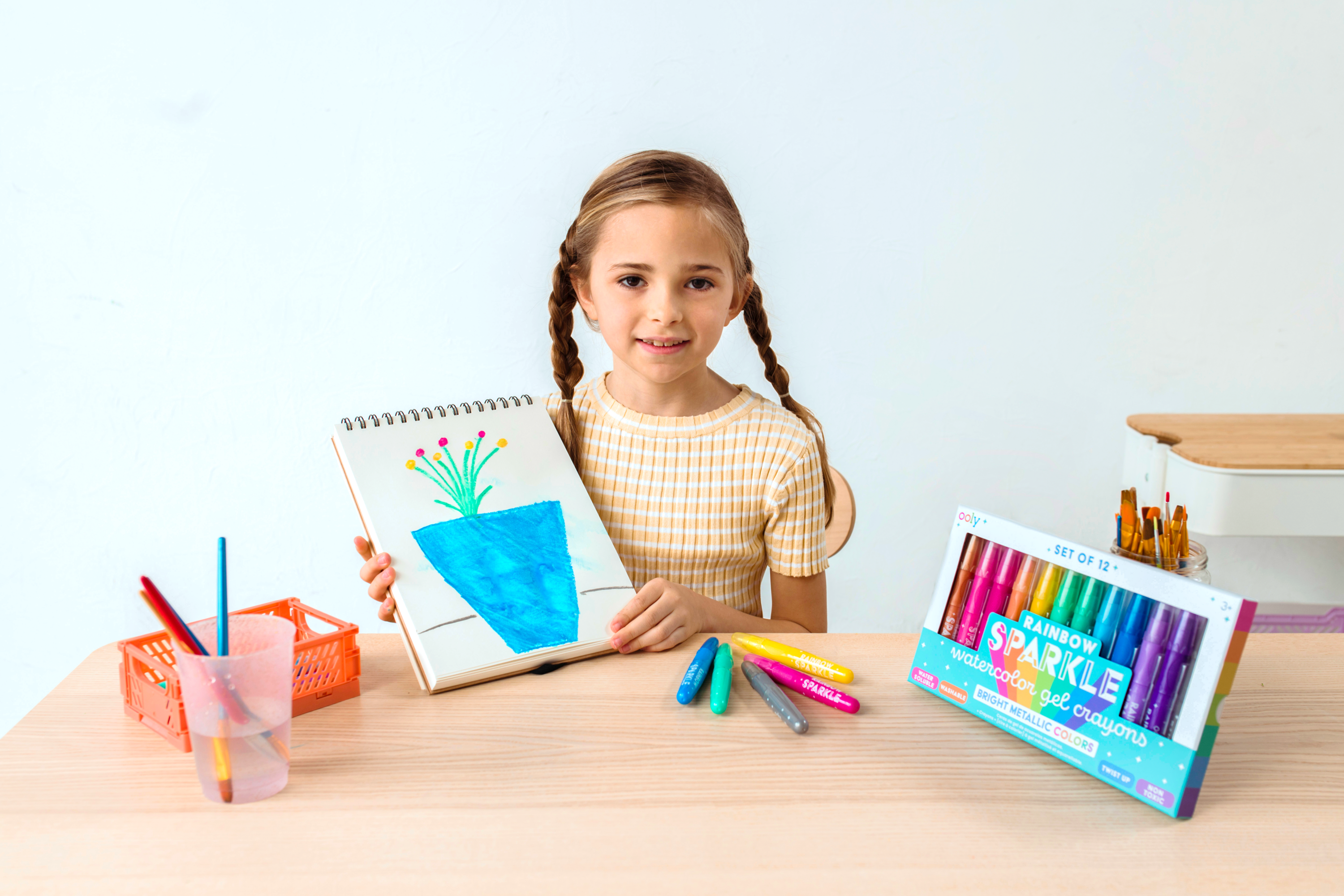 Girl holding completed coloring of blue flower pot with OOLY Rainbow Sparkle Metallic Watercolor Gel Crayons