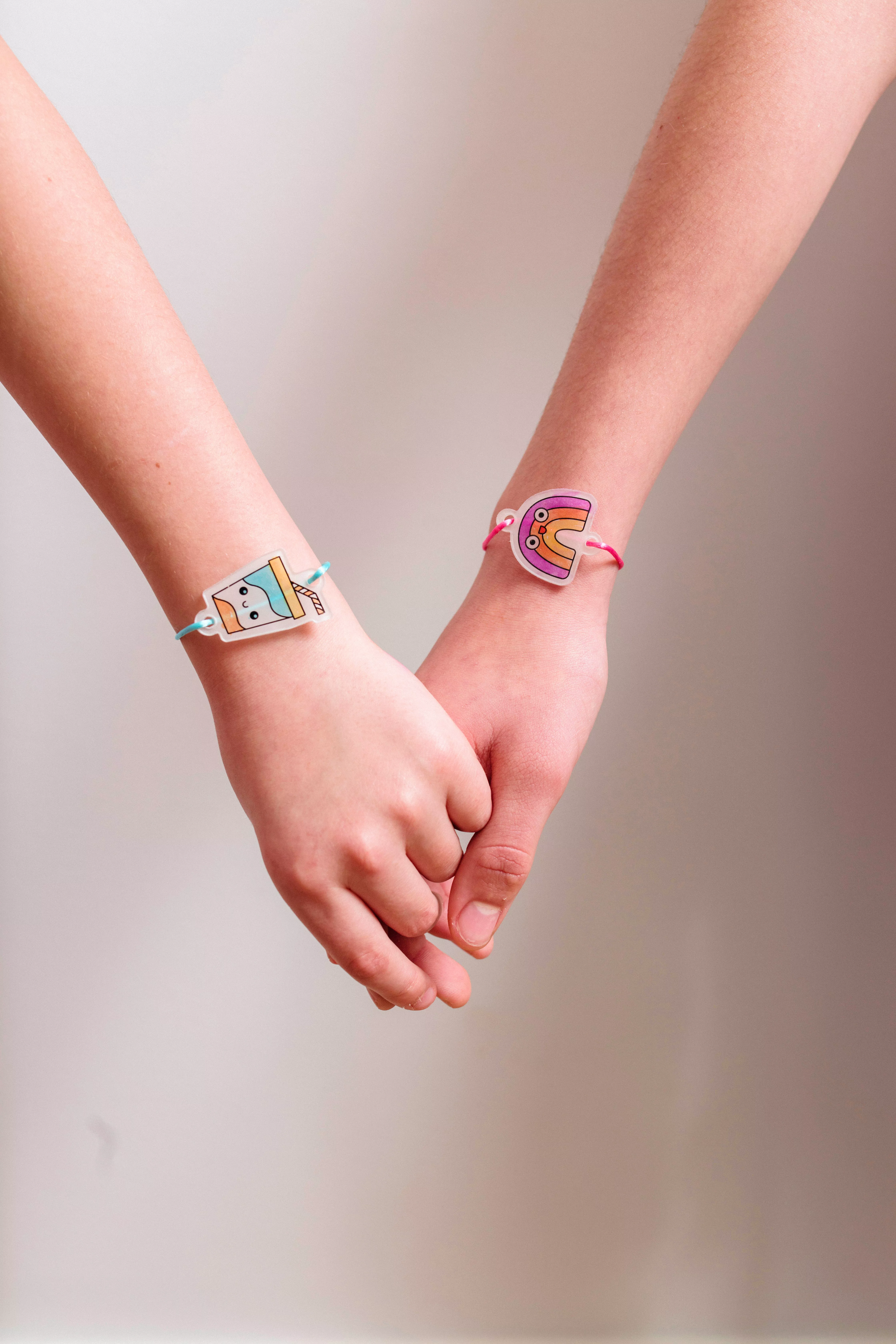 Two hands holding with finished milk shake and rainbow charm bracelets from OOLY Shrink-its! DIY Shrink Art Bracelet Kit - BFF