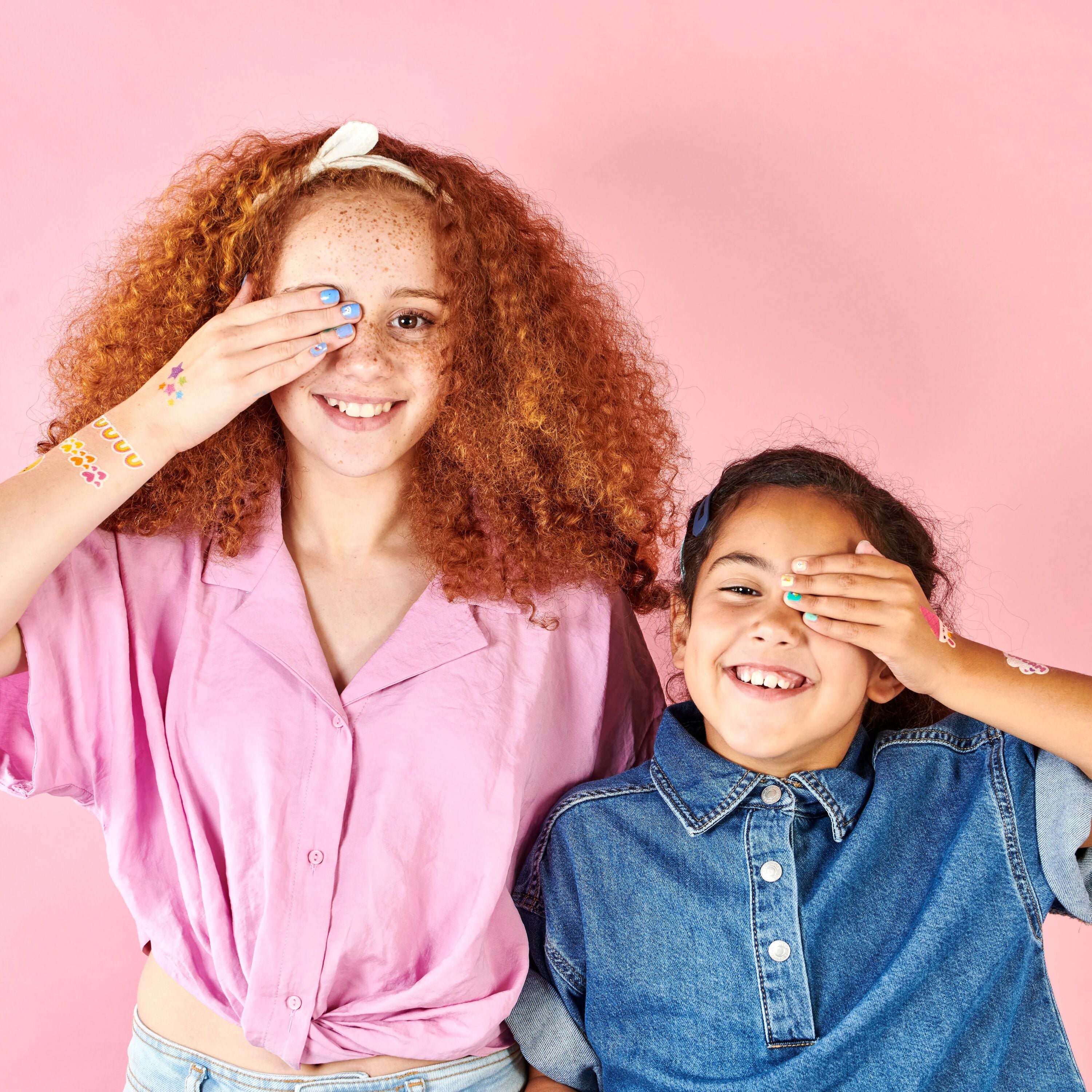 Two girls showing off OOLY Tattoo-Palooza nail stickers and temporary tattoos holding their hands over their eyes with a pink background