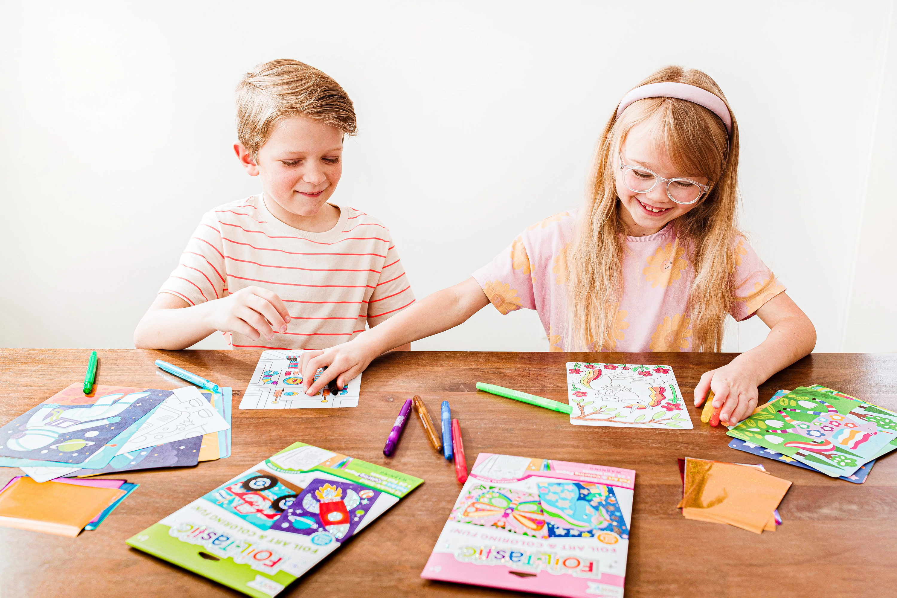 Two kids playing with OOLY Foil-tastic Foil Art Coloring Sets on wooden table wide shot